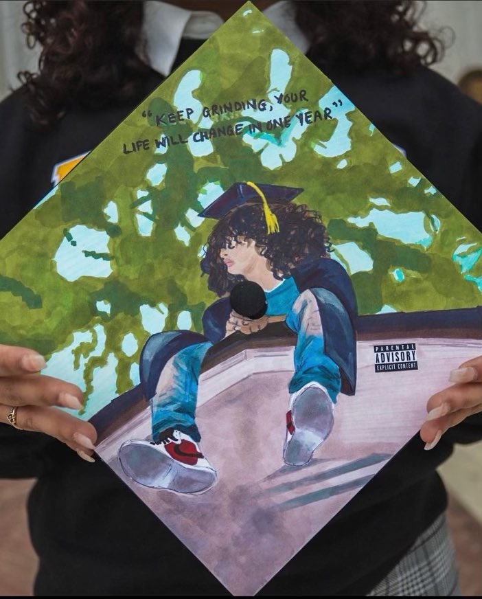 A commencement regalia cap with a design on it similar to a J. Cole album cover - the cap has a student sitting on the edge of a building in jeans and Air Force Ones with a tree in the background.