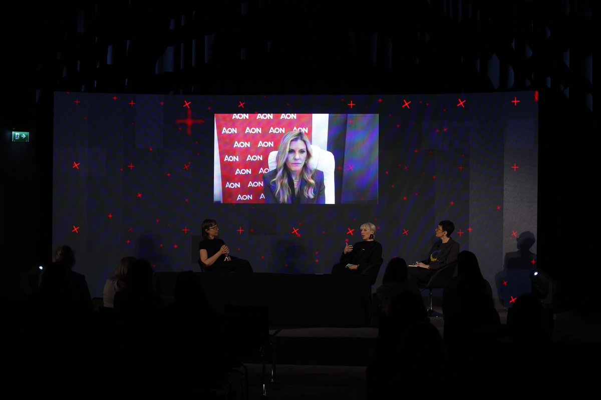 A panel on stage with three in-person speakers and one speaker dialling in remotely on the screen behind the stage. 