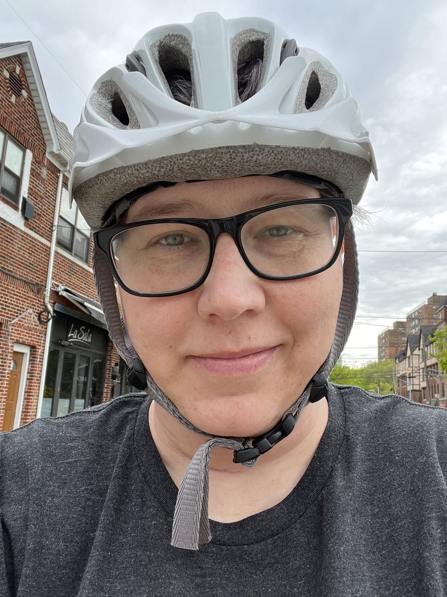A woman in a grey t-shirt and white bike helmet smiles at the camera in front of a grey NYC sky.