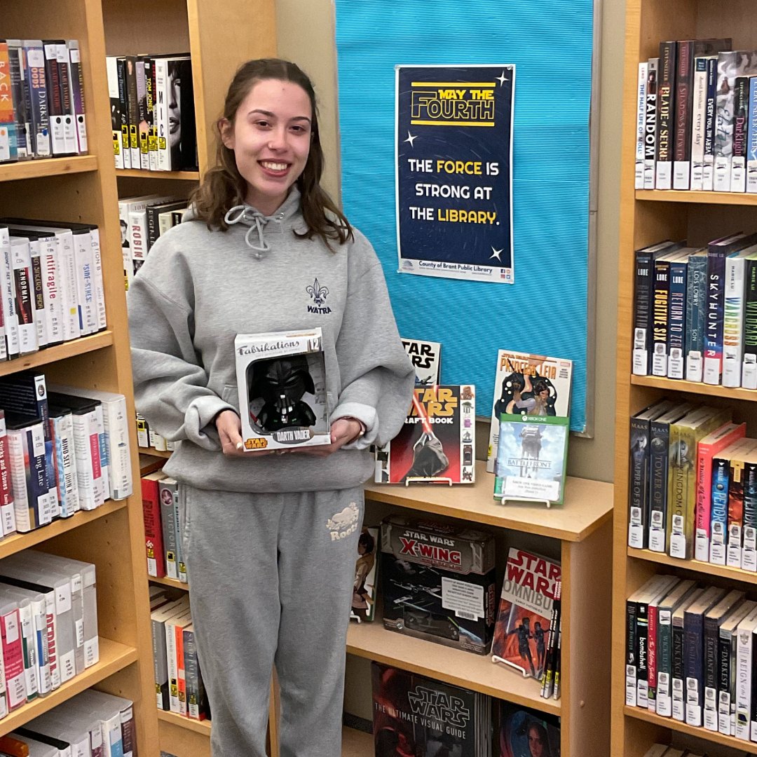 Young girl holding Darth Vader bobblehead, standing in front of a Star Wars book display in a library setting.