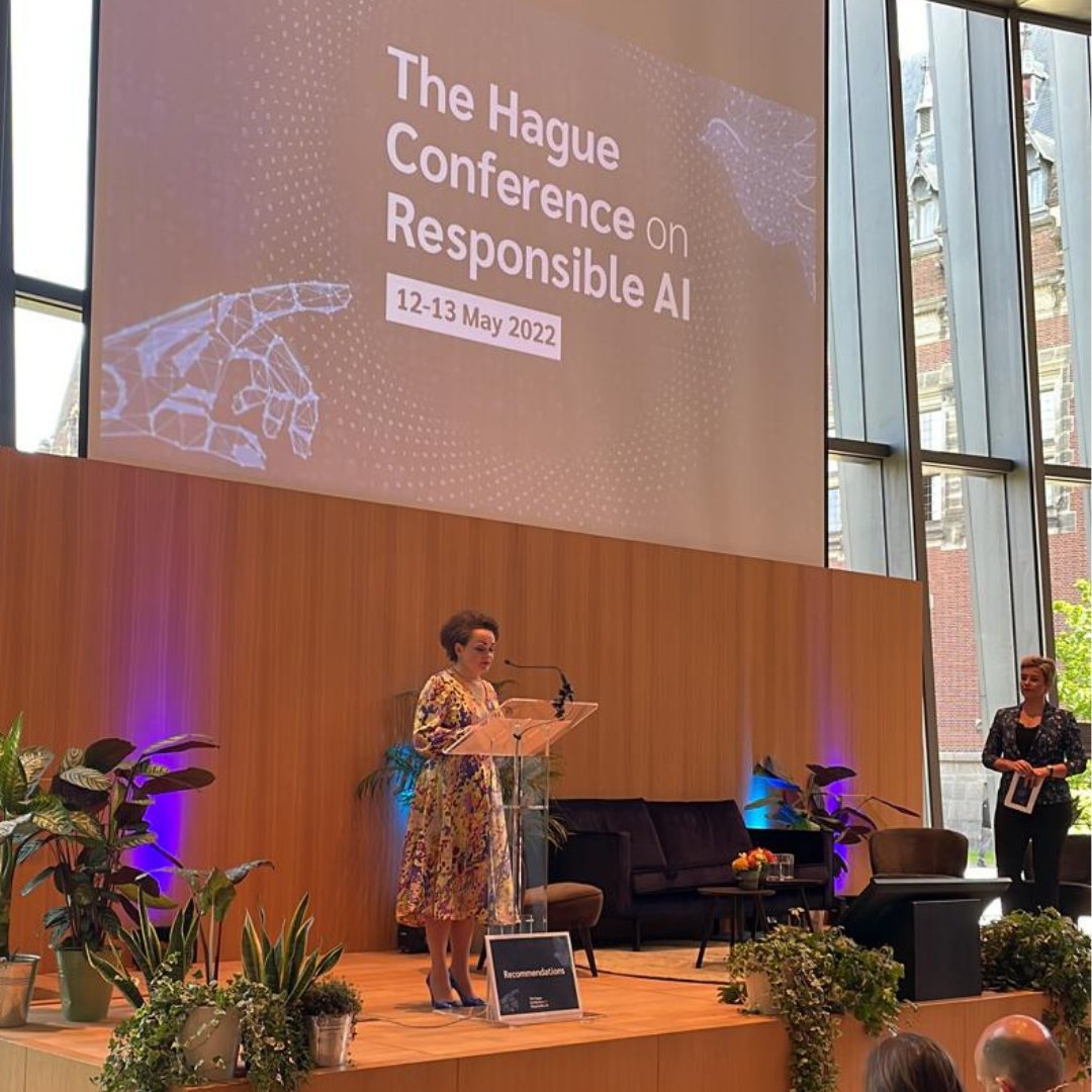 Alexandra van Huffelen staat op het podium achter een spreekgestoelte. Achter haar op een groot scherm staat The Hague Conference on Responsible AI, 12 13 May 2022. Op de achtergrond zijn de hoge ramen te zien.
