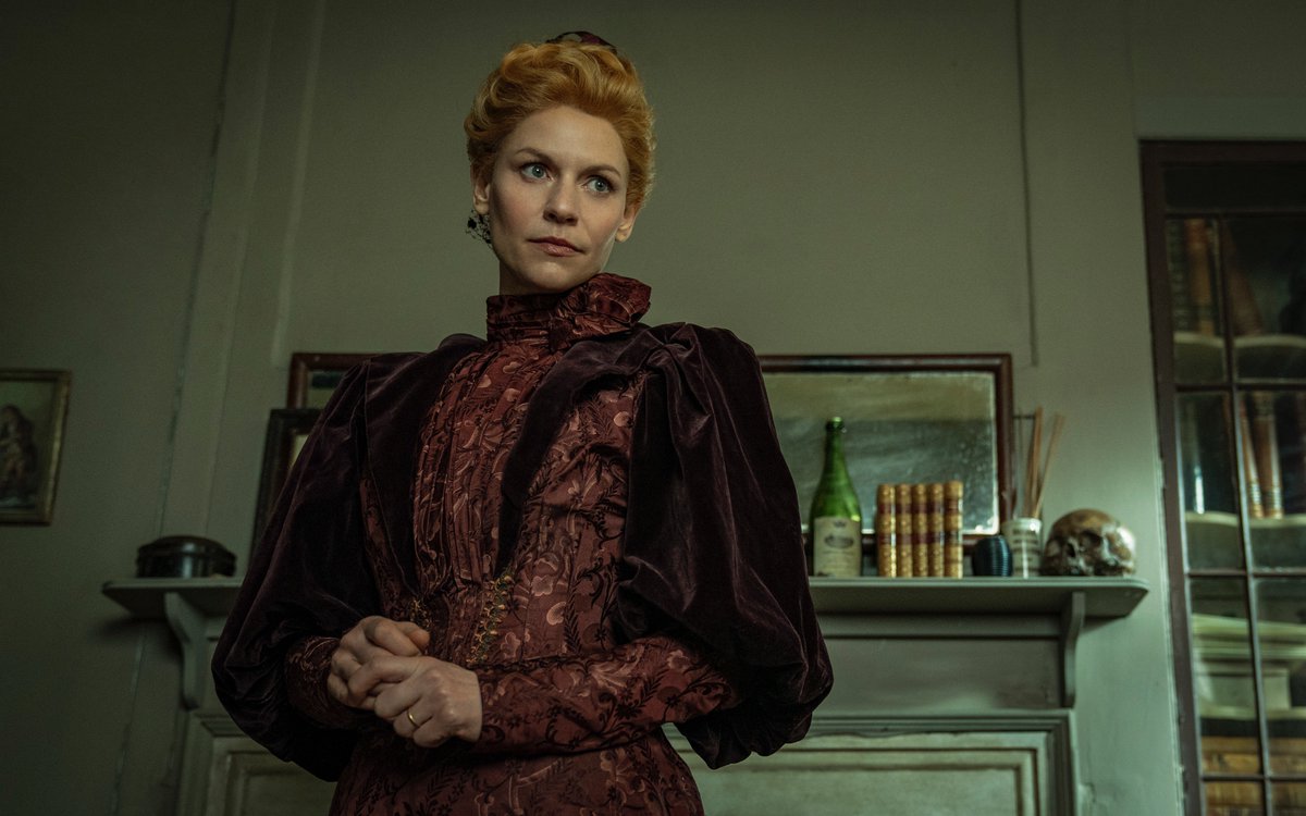 Cora Seaborne (Claire Danes) stands in an ornate red dress with her hair in a structured updo in a home in The Essex Serpent.