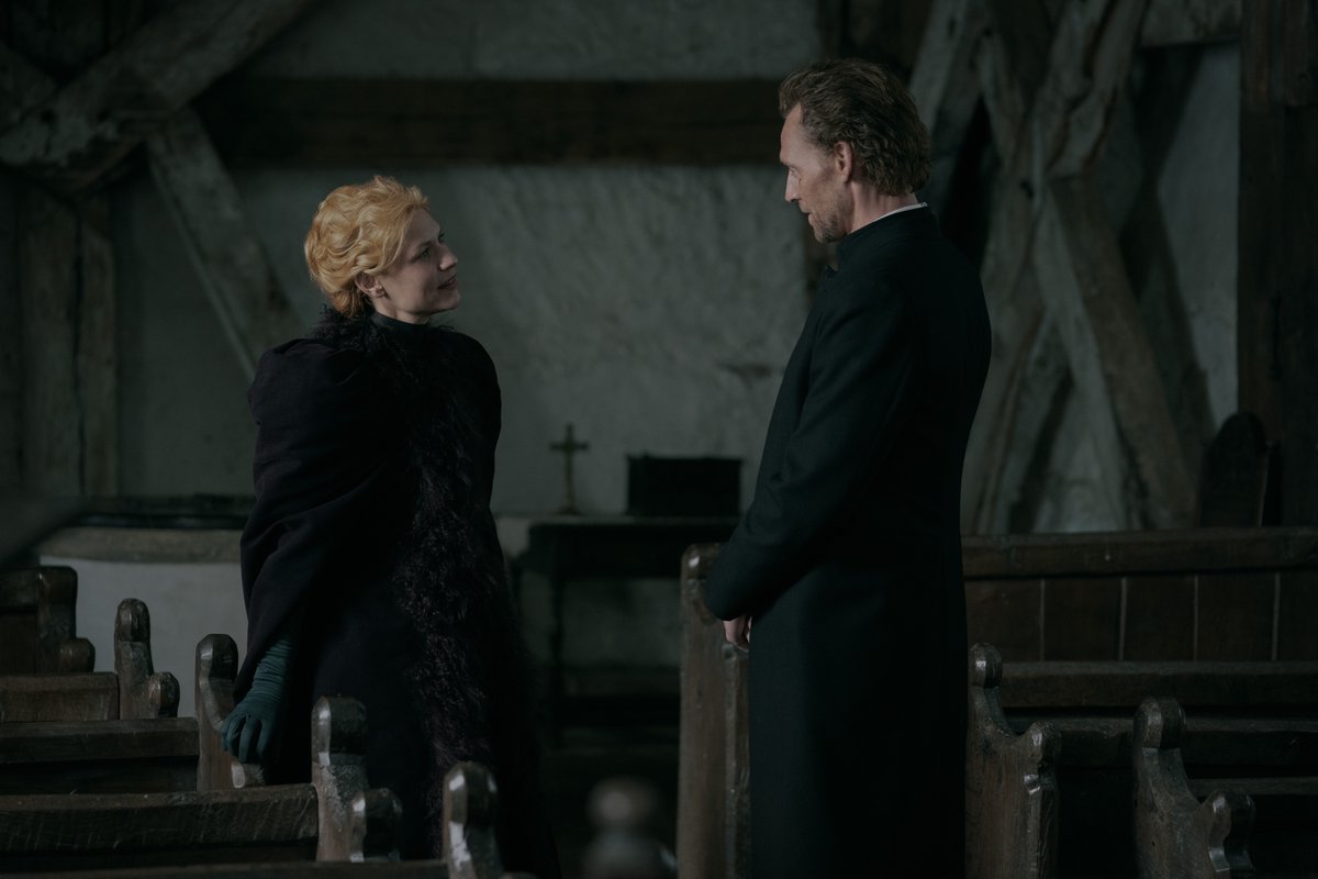 Cora Seaborne (Claire Danes) and Will Ransome (Tom Hiddleston) from The Essex Serpent stand opposite each other in Will’s church, wearing dark clothing.
