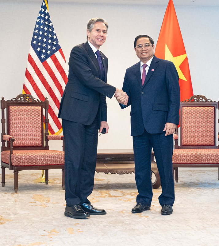 Secretary of State Antony Blinken shakes hands with Vietnamese Prime Minister Chinh, both standing in front of two ornamental wood and rose-colored chairs with the United States and Vietnam flags in the background.