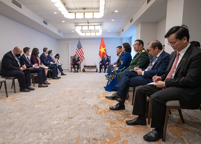 In a long room with U.S. and Vietnamese flags sit U.S. and Vietnamese officials, listening and watching U.S. Secretary of State ANTONY BLINKEN sit and talk with Vietnamese Prime Minister Chinh in chairs at the end of the room.
