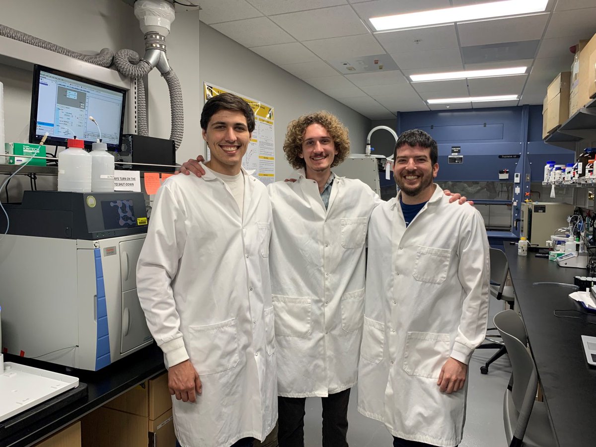 Photo of three white men in lab jackets with their hands around each other's shoulders, standing in a chemistry lab