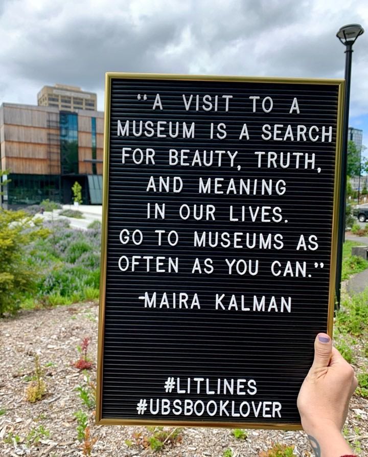 A black letterboard with white letters and gold frame held outside in front of the Burke Museum in Seattle, WA: "A visit to a museum is a search for beauty, truth, and meaning in our lives. Go to museums as often as you can." -Maira Kalman #LitLines #UBSBookLover