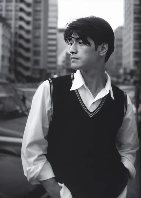 A black and white photograph of Takeshi Kaneshiro. He wears a sweater vest and casually styled dress shirt. He gives a thoughtful look off-camera while a cityscape towers behind him.