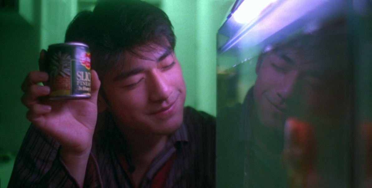 A film still from CHUNGKING EXPRESS. Takeshi Kaneshiro smiles while holding up a can of sliced pineapple to a tank of fish.