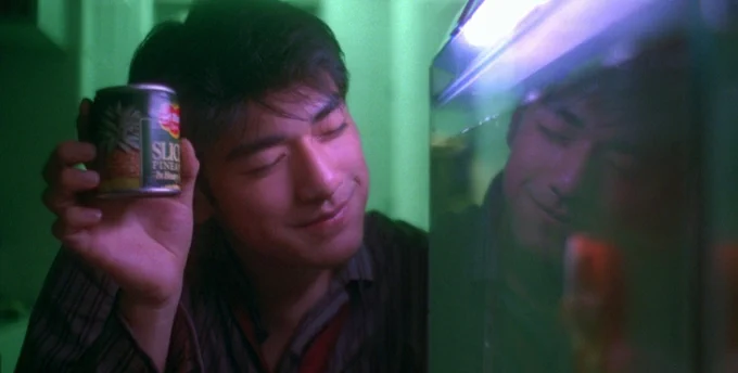 A film still from CHUNGKING EXPRESS. Takeshi Kaneshiro smiles while holding up a can of sliced pineapple to a tank of fish.