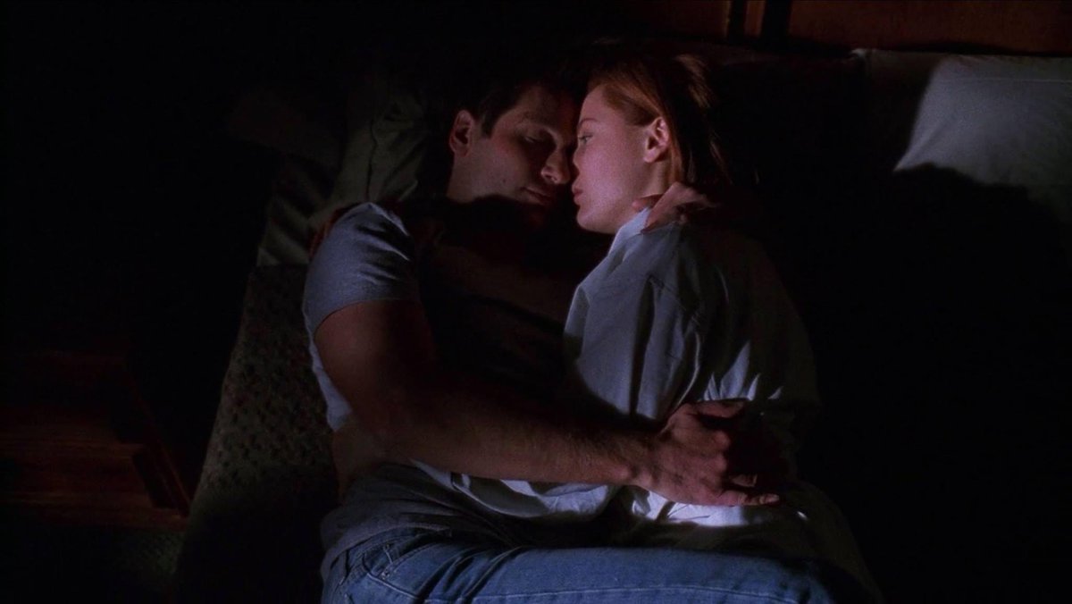 Mulder, tall man with dark brown hair wearing gray T-shirt and jeans hugging Scully, short red haired woman wearing white bathrobe, on a bed in a dark room. From The X-Files original series finale episode The Truth, May 19 2002z