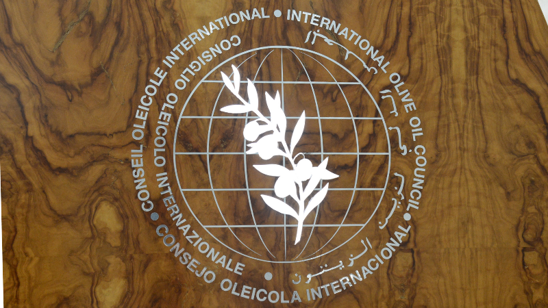 Logotipo del Consejo Oleícola Internacional (COI) que consiste en una rama de olivo sobre el globo terráqueo rodeado del nombre de la entidad. Todo ello sobre madera de olivo. 