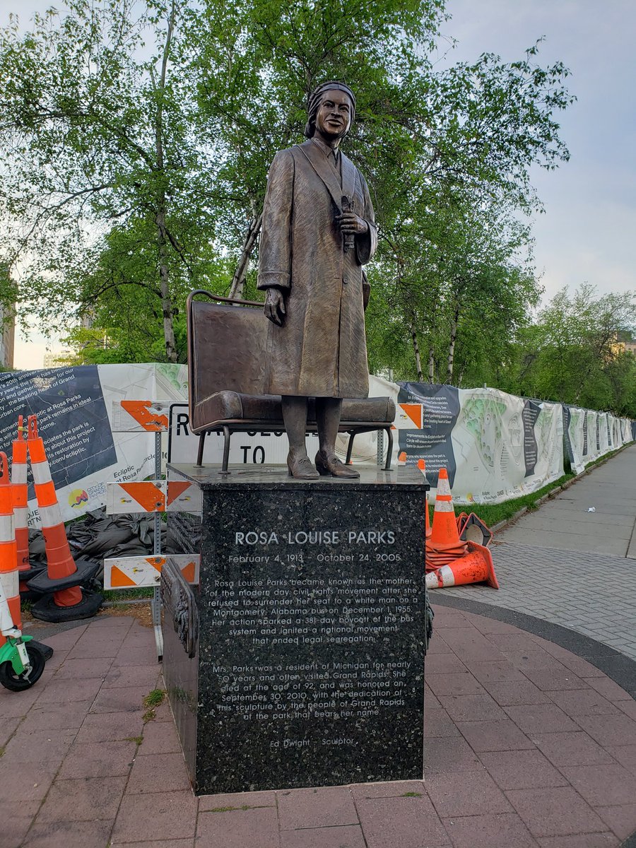 A bronze statue of Rosa Parks. Behind her are the trees of Rosa Parks Circle. Parks stands in front of a bronze bus seat. She holds a pair of gloves and has a purse over her left shoulder.