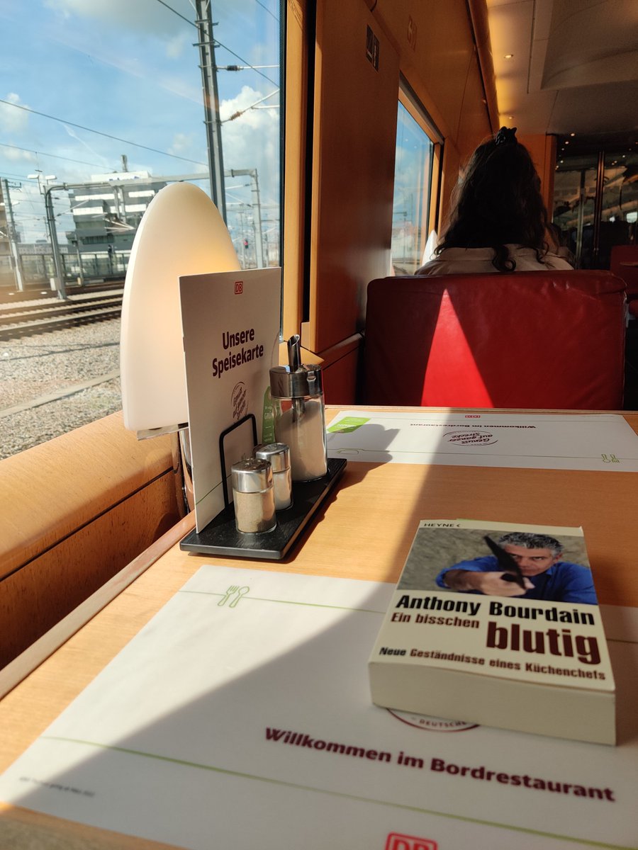 View from the table in a dining Car onboard an Intercity Express. On the left, the trainwindow with a blue sky in the background. . Showing the table with a book from Anthony Bourdain "Ein bisschen blutig" 
