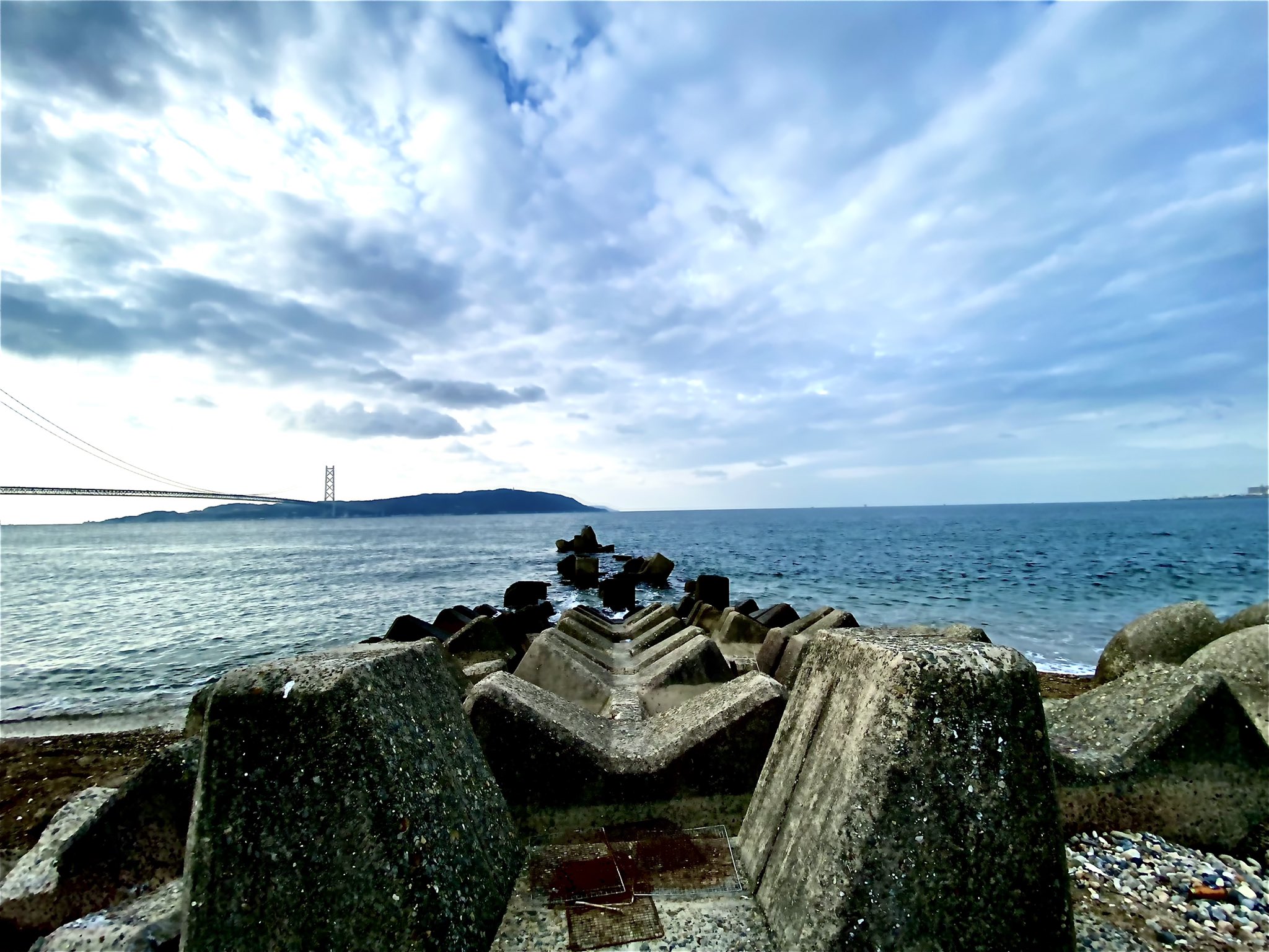 丸い三角 消波ブロック越しに海を 彼方に明石海峡大橋 休日は徒歩や自転車で海辺を散策のがルーティン 海 海岸 海辺 消波ブロック コンクリート二次製品 護岸 散歩 空 雲 明石海峡 瀬戸内海 明石海峡大橋 写真 写メ Photograph