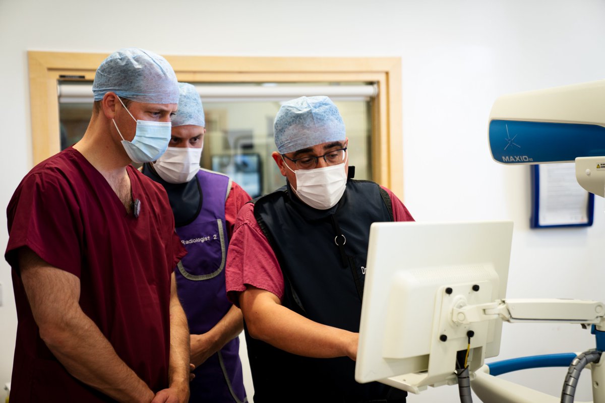 The Duke of Cambridge, standing on the left wearing medical scrubs, is shown a screen during an interventional radiology procedure