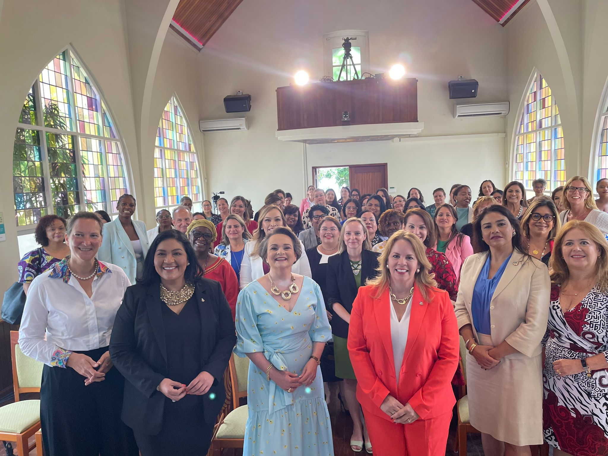 Alexandra van Huffelen en Minister-President Evelyn Wever-Croes van Aruba staan samen met andere vrouwen vooraan een grote groep vrouwen. Iedereen kijkt in de camera. De groep staat in een grote, lichte kerk, met op de achtergrond glas-in-lood ramen. 