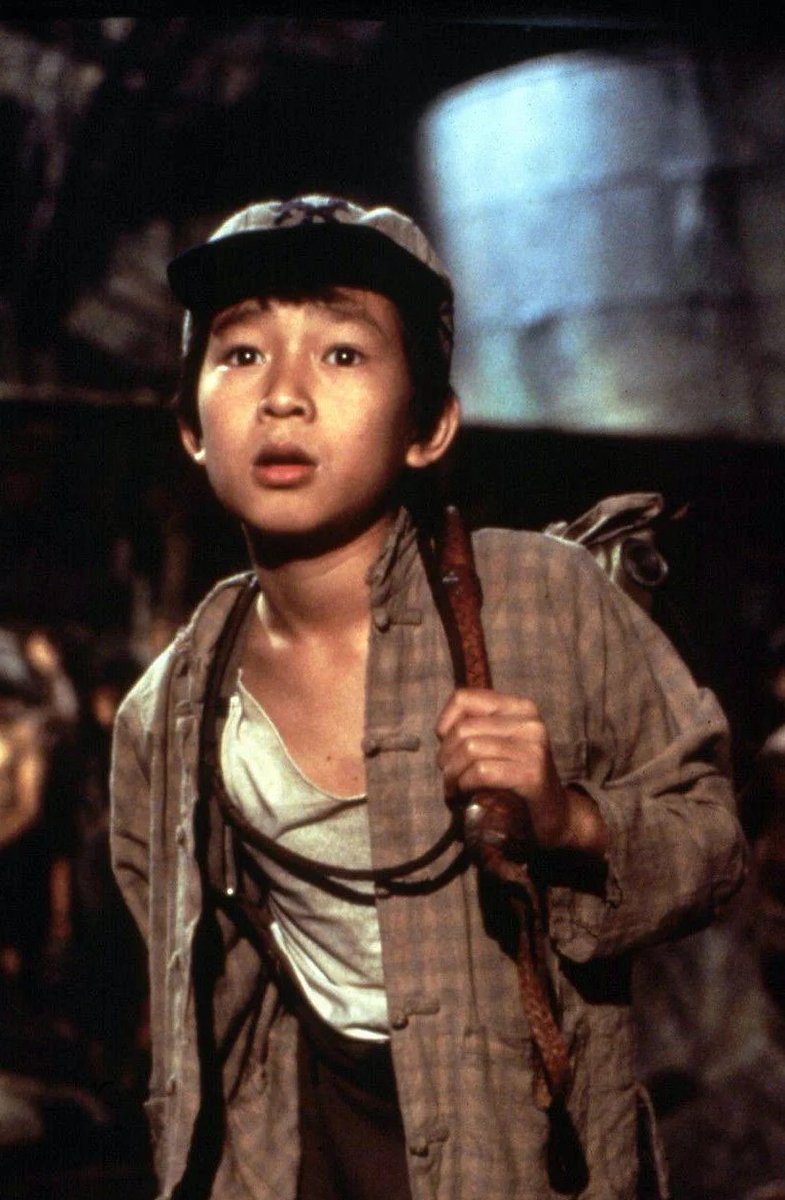 A photograph of a young Ke Huy Quan on the set of INDIANA JONES AND THE TEMPLE OF DOOM. He wears a baseball cap and carries a whip.