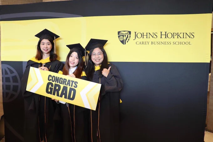 Photo of three students posing with a graduation prop in front of Carey logo wall.
