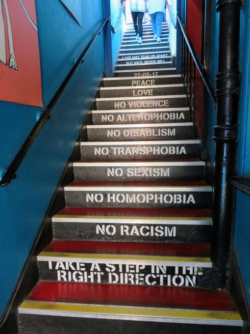Eine schmale Treppe in schwarz, gelb, rot, zwischen blauen Wänden. Jede Stufe vermittelt eine Botschaft, z.B. take a step in the right direction, no racism, no homophobia, no sexism, no transphobia......,love, peace... Die Treppe ist von unten fotografiert, ganz oben sieht man zwei Menschen die hoch gehen.