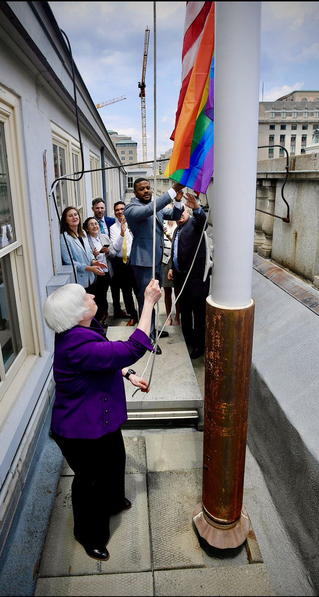 Secretary Yellen pulls a rope to raise the Pride flag, which is seen underneath the American flag, up a flagpole. A small group of people are seen behind her cheering.