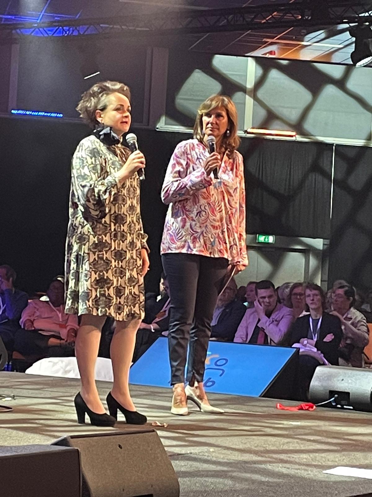 Alexandra van Huffelen staat op een podium naast Astrid Joosten. Beiden hebben een microfoon in de hand. 