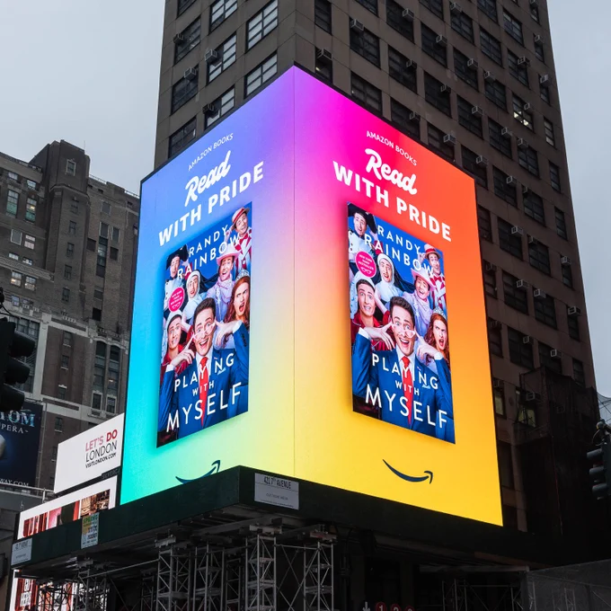 A billboard with a rainbow gradient background, featuring an image of the book "Playing With Myself". The cover includes several overlapping images of Randy Rainbow in different costumes. The text on top reads "Read With Pride".