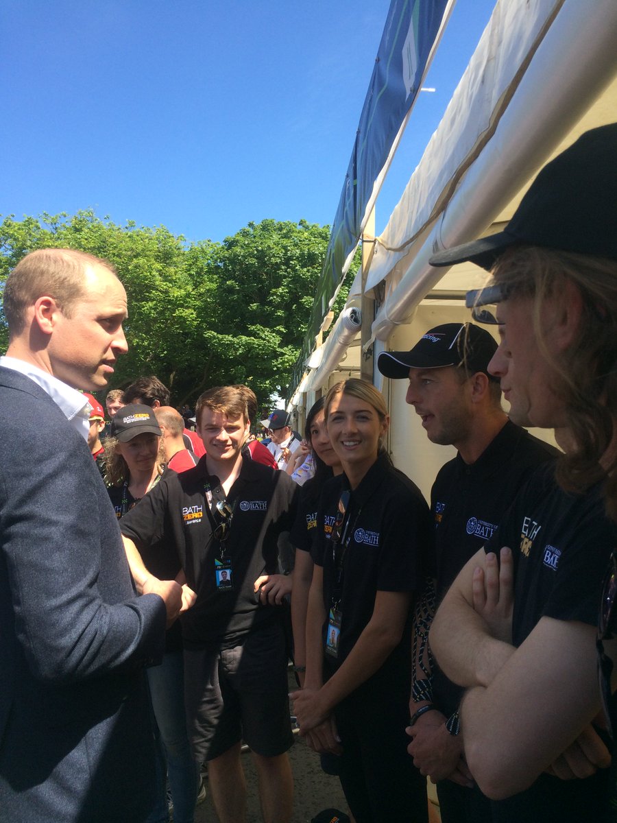 Prince William talking with several Bath students. The students are wearing black uniforms with several wearing matching baseball caps. The weather is sunny and the sky is blue and cloudless.