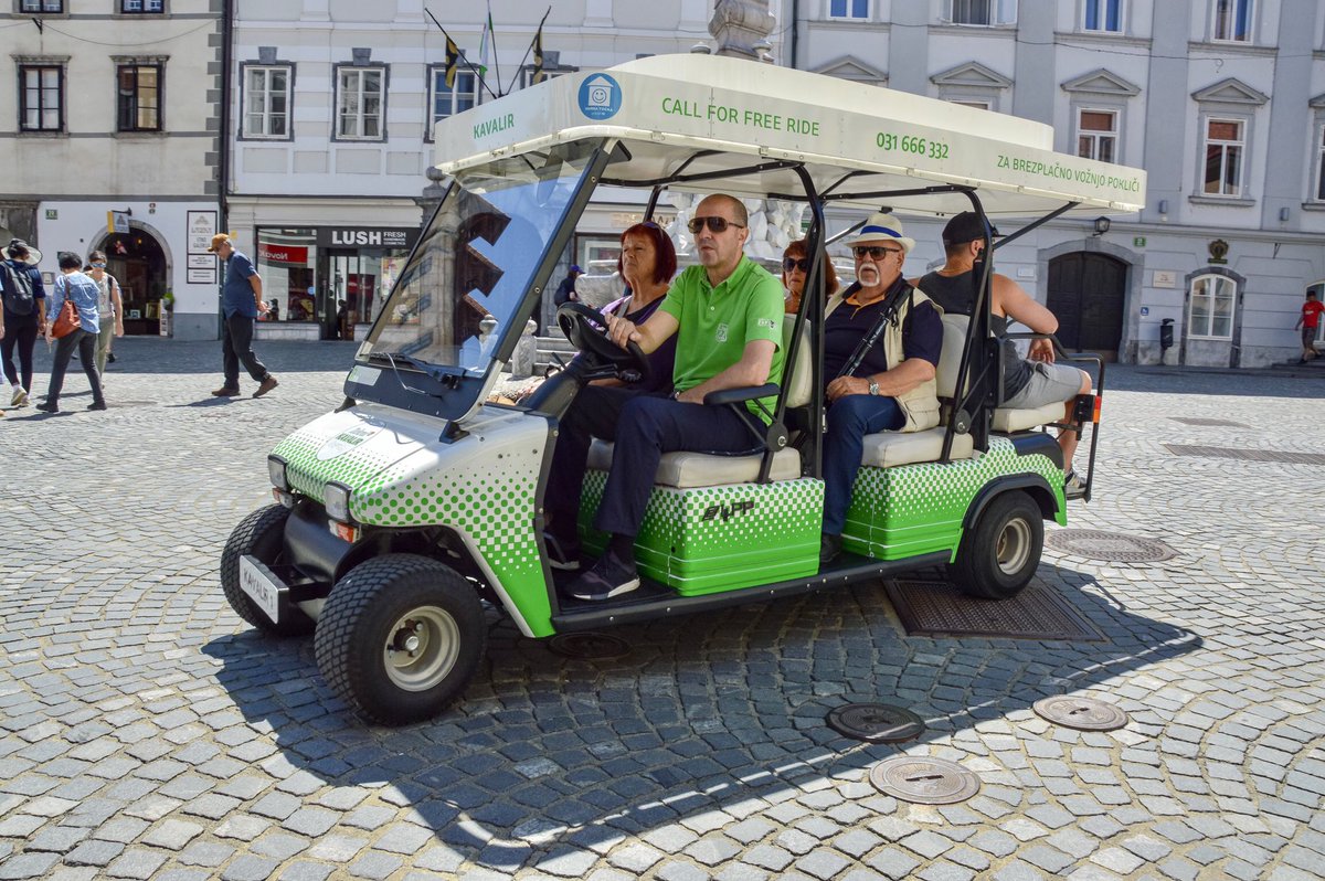 A green-and-white light electric vehicle moves through a European city. It holds five passengers, including a man with a cane.