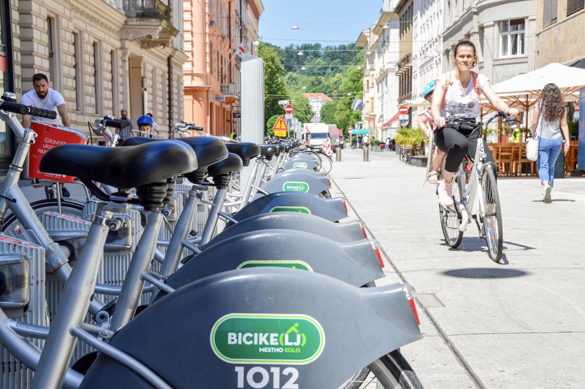 A row of shared bicycles with gray skirt guard and a green logo. A woman cycles by out of focus to the right.
