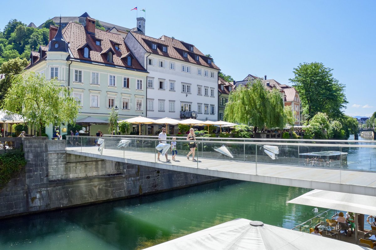 A concrete bridge with a glass railing stretches across green river in a medieval European city.