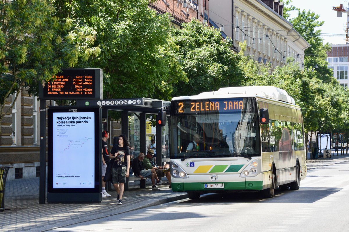 A bus sits at a bus stop with an electronic sign indicating the arrival times. Passengers sit in the glass structure.