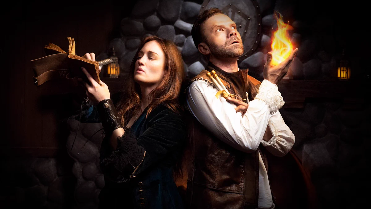 Ashley Johnson writing in a leather-bound notebook & Liam O'Brien with a magical fireball stand back to back in Shakespearean costumes