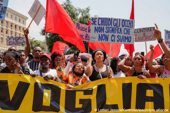 A large group of protesters (most of them Black) are walking down a street, carrying a banner and posters. One of the posters reads (in Italian): "if you close your eyes it doesn't mean we're not there".