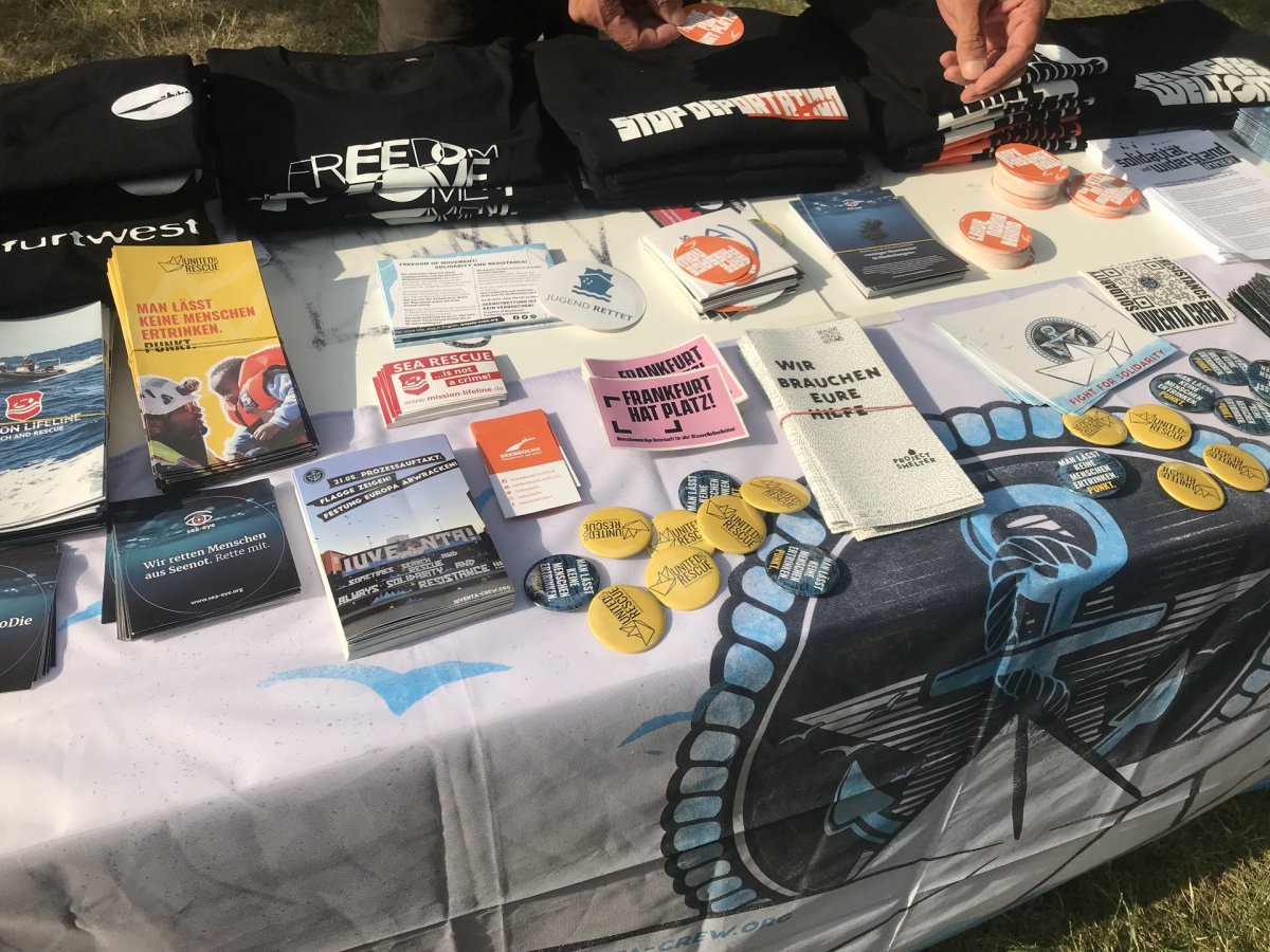 Ein Infotisch.<br>Tischdecke ist die IUVENTA-Crew Soliflagge.<br>Auf dem Tisch sind T-Shirts mit SB Ffm Logo, frankfurt west, FREEDOM OF MOVEMENT, STOP DEPORTATION und mehr.<br>Flyer und Sticker von  Mission Lifeline, United4Rescue, Sea-Eye, IUVENTAA Crew, Frankfurt hat Platz, Project Shelter, Jugend rettet, Sea Watch.