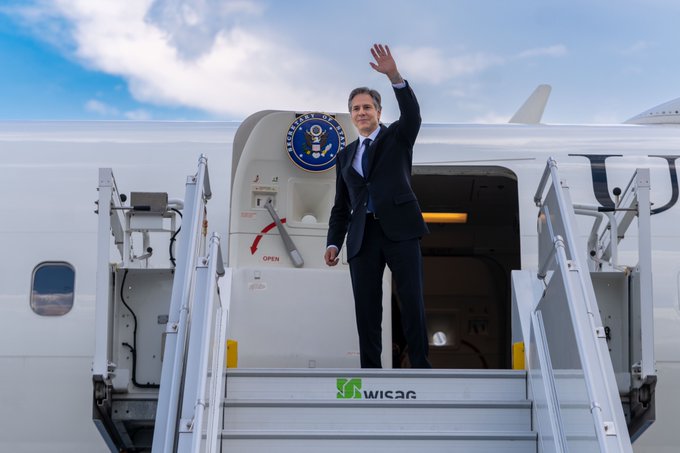 Secretary of State Antony J. Blinken stands outside of the open door of an airplane in dark suit, waving against the backdrop of clear skies. An official Secretary of State seal hangs from the aircraft door.