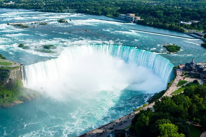 Water flows over the Canadian Niagara Falls (Horseshoe Falls). A power station is visible upriver.
