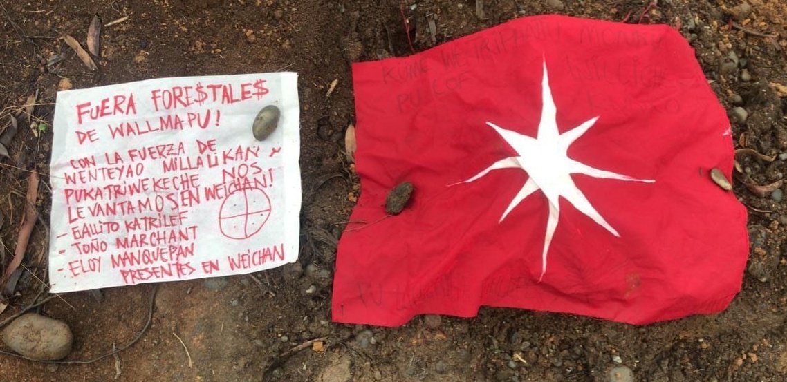 picture of a red mapuche flag & canvas on the floor, it reads : « FUERA FORESTALES DEL WALLMAPU! Con la fuerza de Wenteyao, Millalikan, Pukatriwekeche. Nos levantamos en weichan! - Gallito Katrilef - Toño Marchant - Eloy Manquepan Presentes en Weichan »