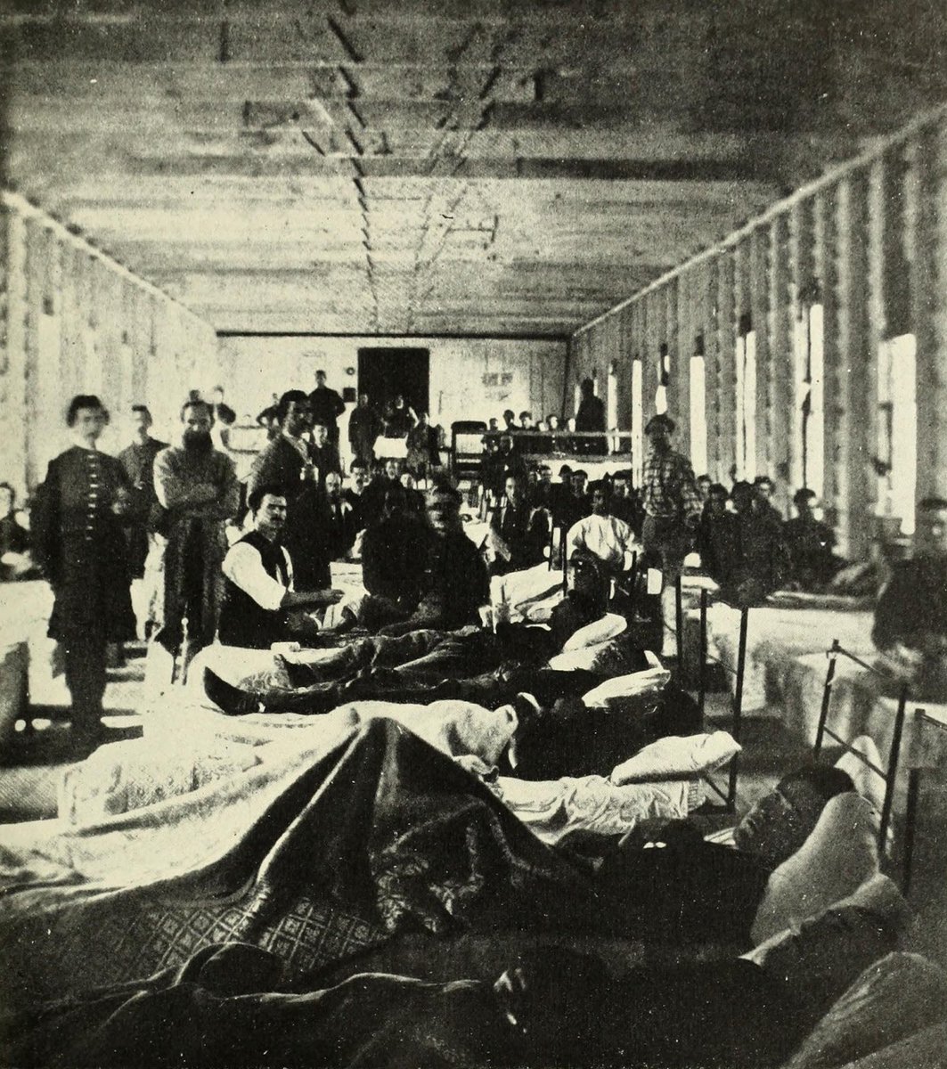 A black and white photograph showing civil war soldiers convalescing in a makeshift hospital. It looks very cramped and grim. There are many men in the image, some are lying in bed, others are walking around.