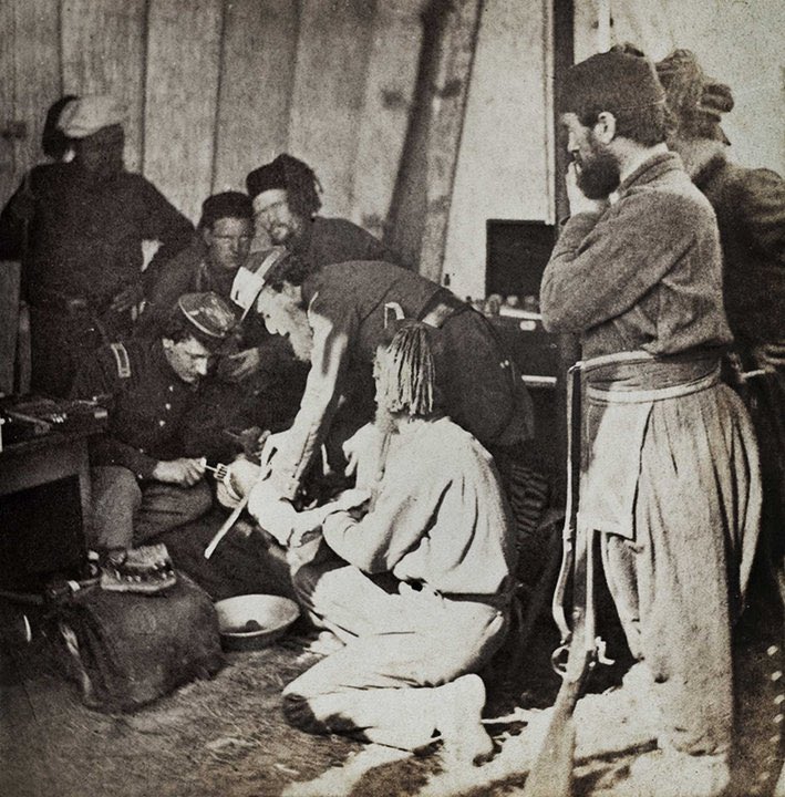 A black and white photo of a civil war field hospital and what looks like an amputation taking place. There are 8 men in the image and they are very cramped in, which gives the image a chaotic feel. Two men look on as one man (presumably the medic) is sawing the leg off another man who is being held down by 2 others. There are two men assisting the doctor. It’s a gruesome, disturbing image.