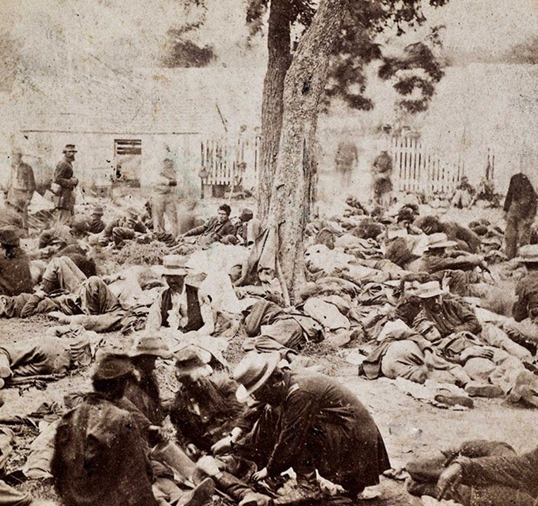 A black and white image of civil war soldiers, lying around outside, after a battle. It looks like piles of bodies , but they are alive. Most are lying down, but there are some walking about or checking on the injured.