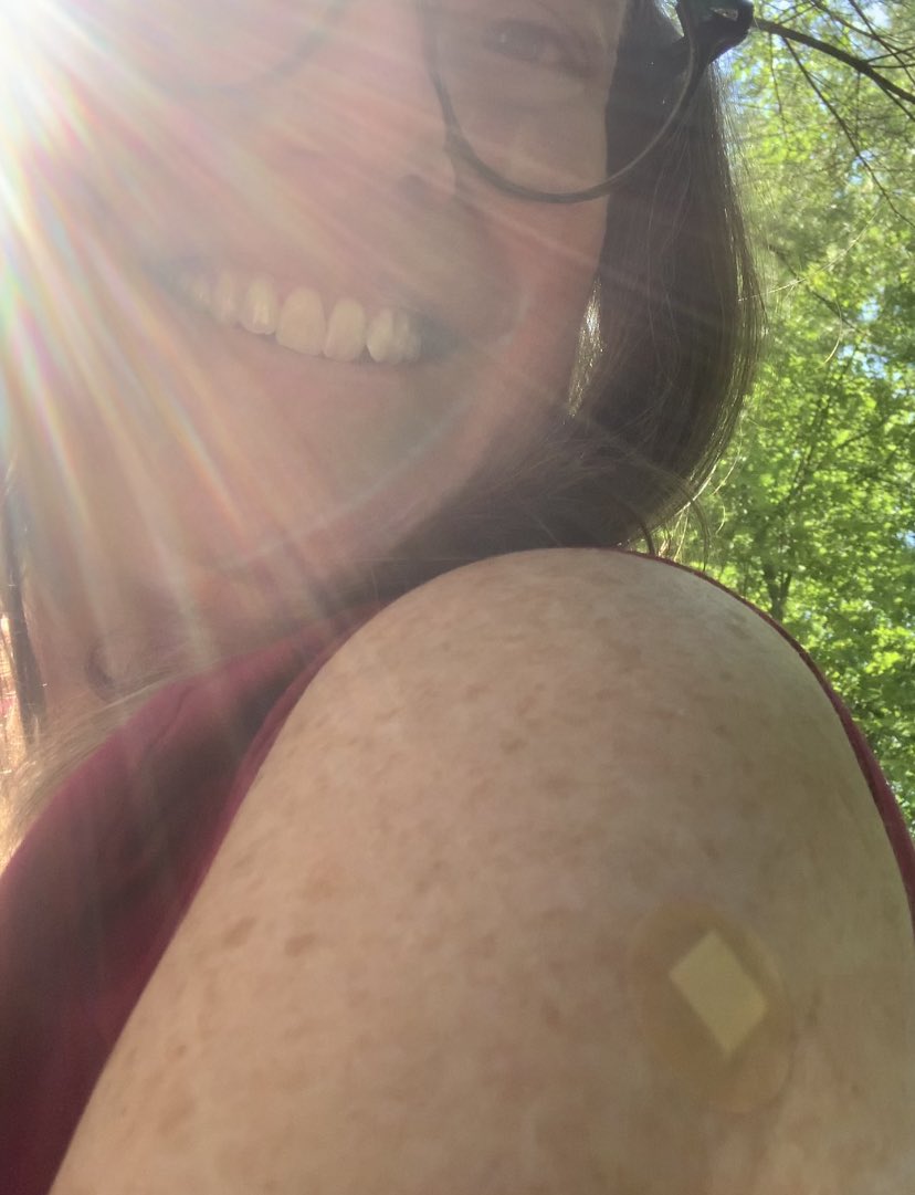 White woman with freckles showing off small round bandaid on her shoulder.