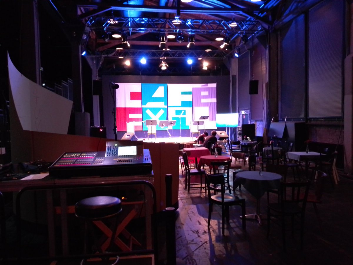 Vorne links ein Sound- & Lichtsteuerungspodium<br>Tischchen mit Stühlen.<br>Ganz hinten leuchtet bunt die Café Exil Bühne.