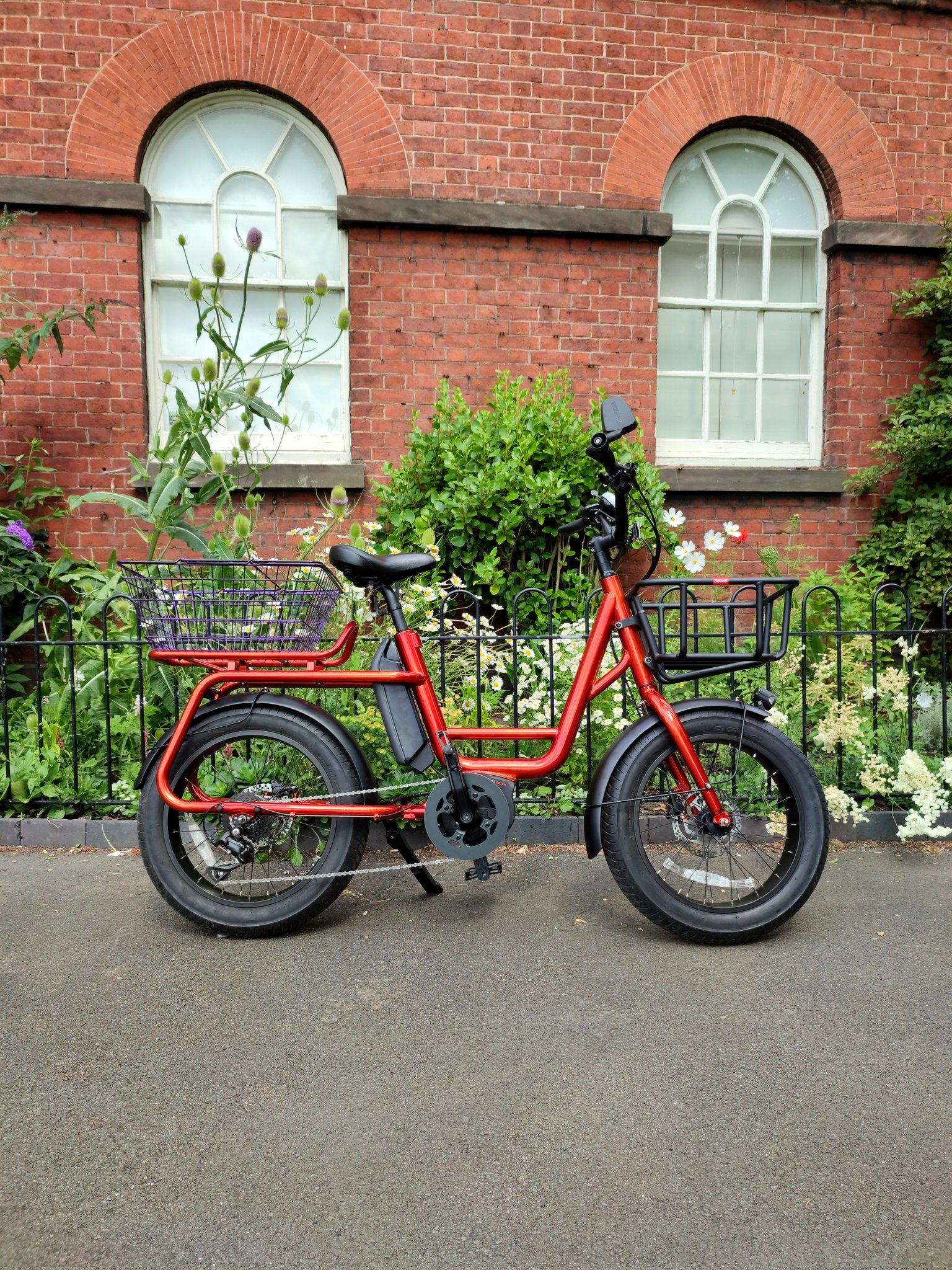 A red electric cargo bike with a purple rear basket and black front basket is in front of a wall, with arch windows and lots of greenery. There are daisies and various flowers amongst the greenery. This view is from the side