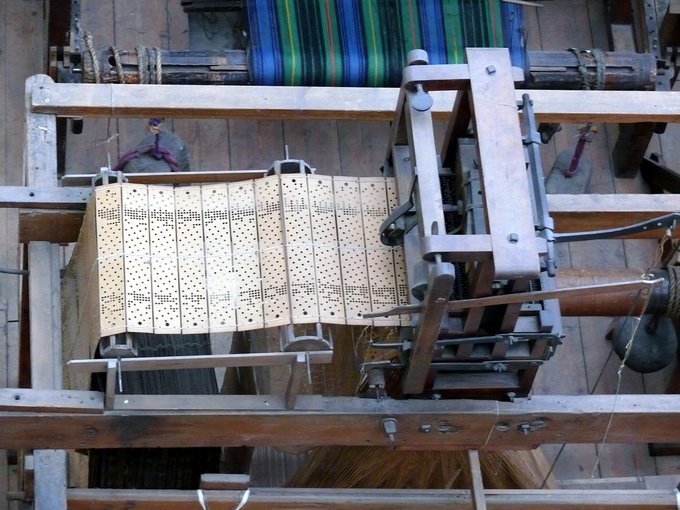 Picture of a set of linked punch cards used to program a Jacquard Loom. The cards are long, skinny rectangles linked at the top and bottom and fed into a metal reader. 
