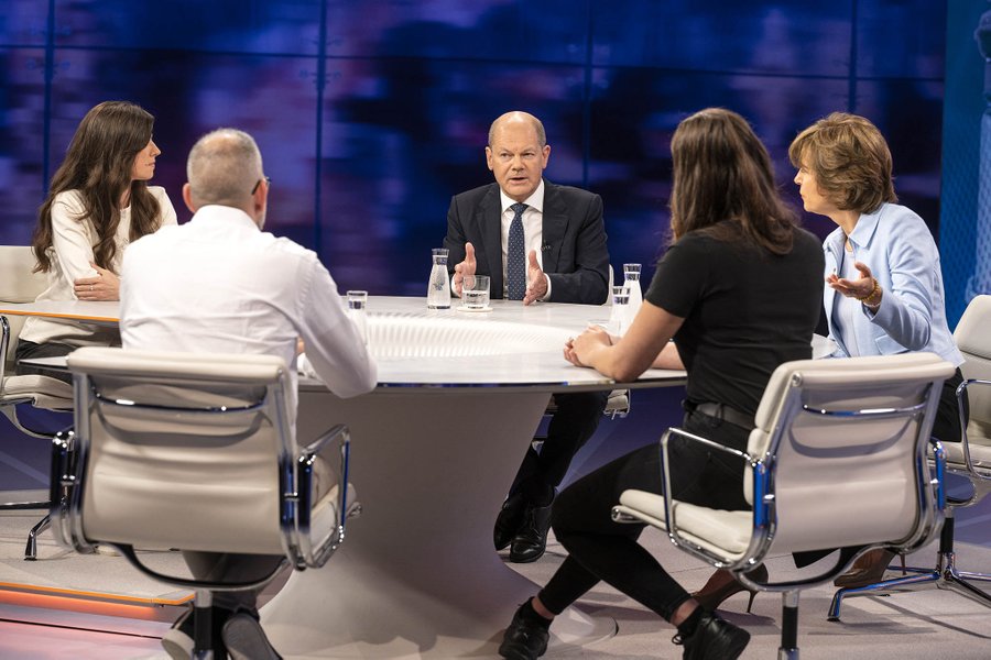 Bundeskanzler Scholz ist im fernsehstudio der talk-Sendung Maybrit Illner zu sehen. Er sitz an einem runden Tisch, im Bild rechts neben ihm die Moderatorin Maybrit Illner. Neben ihm ist die ukrainische velergein Kateryna Msihchenko zu erkennen. Mit den Rücken zur Kamera sitzen  gegenüber dem Bundeskanzler die Studentin Rifka Lambrecht und der Intensivpfleger Ralf Berning.