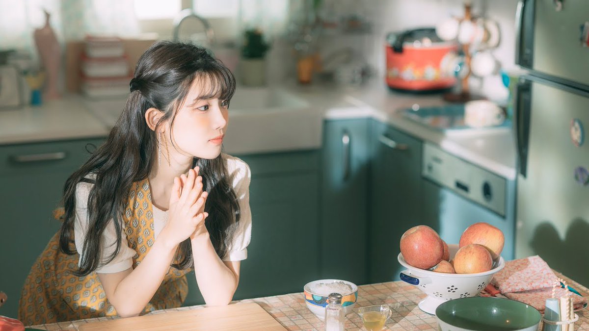 A young woman with long dark hair and bangs sits at a kitchen table with her hands clasped together, wearing a yellow patterned dress over a white blouse, with a bowl of peaches on the table beside her.