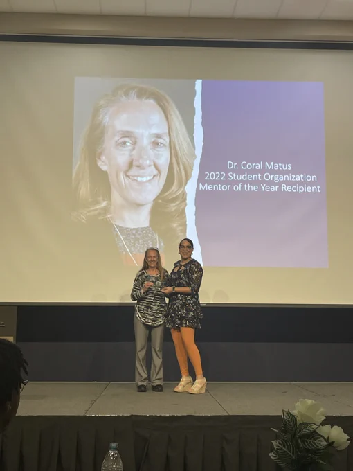 Dr. Coral Matus accepts her Student Organization Advisor of the Year award on stage with her name and photo on the screen behind her.