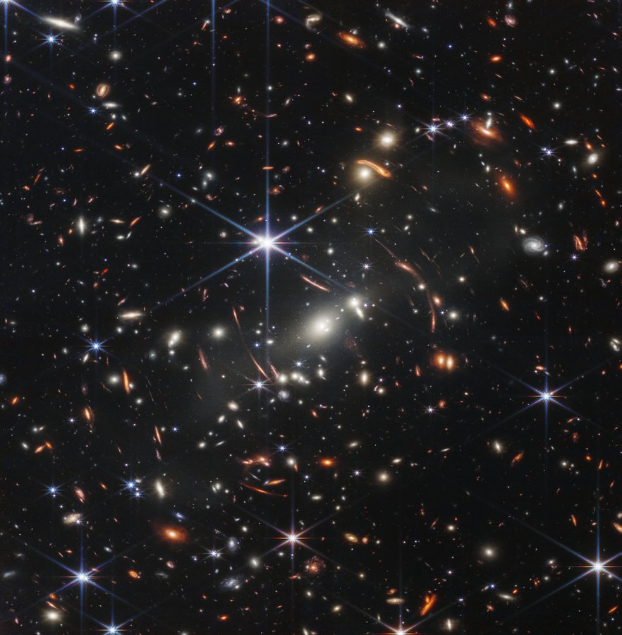 The background of space is black. Thousands of galaxies appear all across the view. Their shapes and colors vary. Some are various shades of orange, others are white. Most stars appear blue, and are sometimes as large as more distant galaxies that appear next to them. A very bright star is just above and left of center. It has eight bright blue, long diffraction spikes. Between 4 o’clock and 6 o’clock in its spikes are several very bright galaxies. A group of three are in the middle, and two are closer to 4 o’clock. These galaxies are part of the galaxy cluster SMACS 0723, and they are warping the appearances of galaxies seen around them. Long orange arcs appear at left and right toward the center.