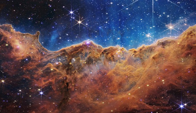Nebulosa de Carina, con un campo estelar que muestra innumerables estrellas de muchos tamaños. Las más pequeñas son puntos de luz pequeños, distantes y débiles. Las más grandes aparecen más grandes, más cercanas, más brillantes y más resueltas con picos de difracción de 8 puntos. La parte superior de la imagen es azulada, y tiene vetas difusas y translúcidas similares a las de las nubes que se elevan desde la nebulosa inferior. La formación nubosa anaranjada de la mitad inferior varía en densidad y va de translúcida a opaca. Las estrellas varían de color, la mayoría tienen una tonalidad azul o naranja. La estructura nubosa de la nebulosa contiene crestas, picos y valles, un aspecto muy similar al de una cordillera. 